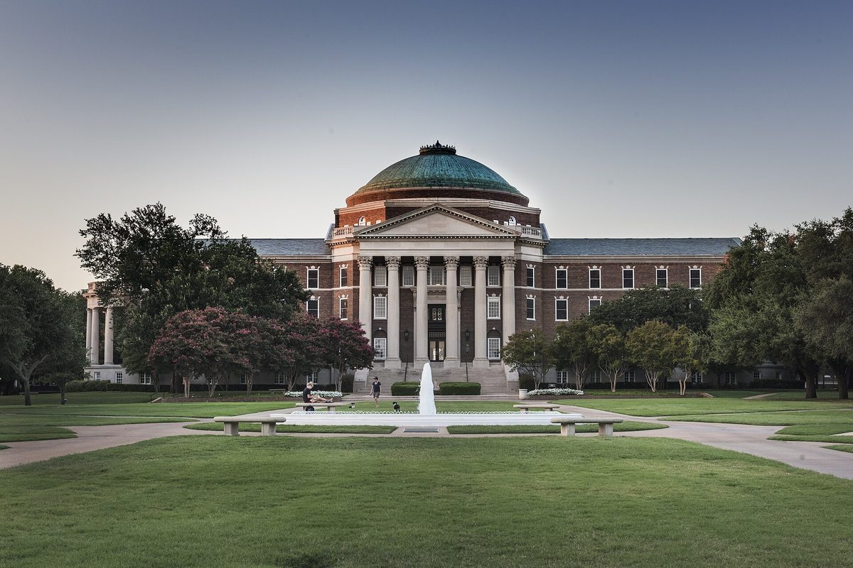 Dallas Hall at Southern Methodist University, a red-brick building with white columns and a green copper dome, behind a fountain and a wide lawn at dusk