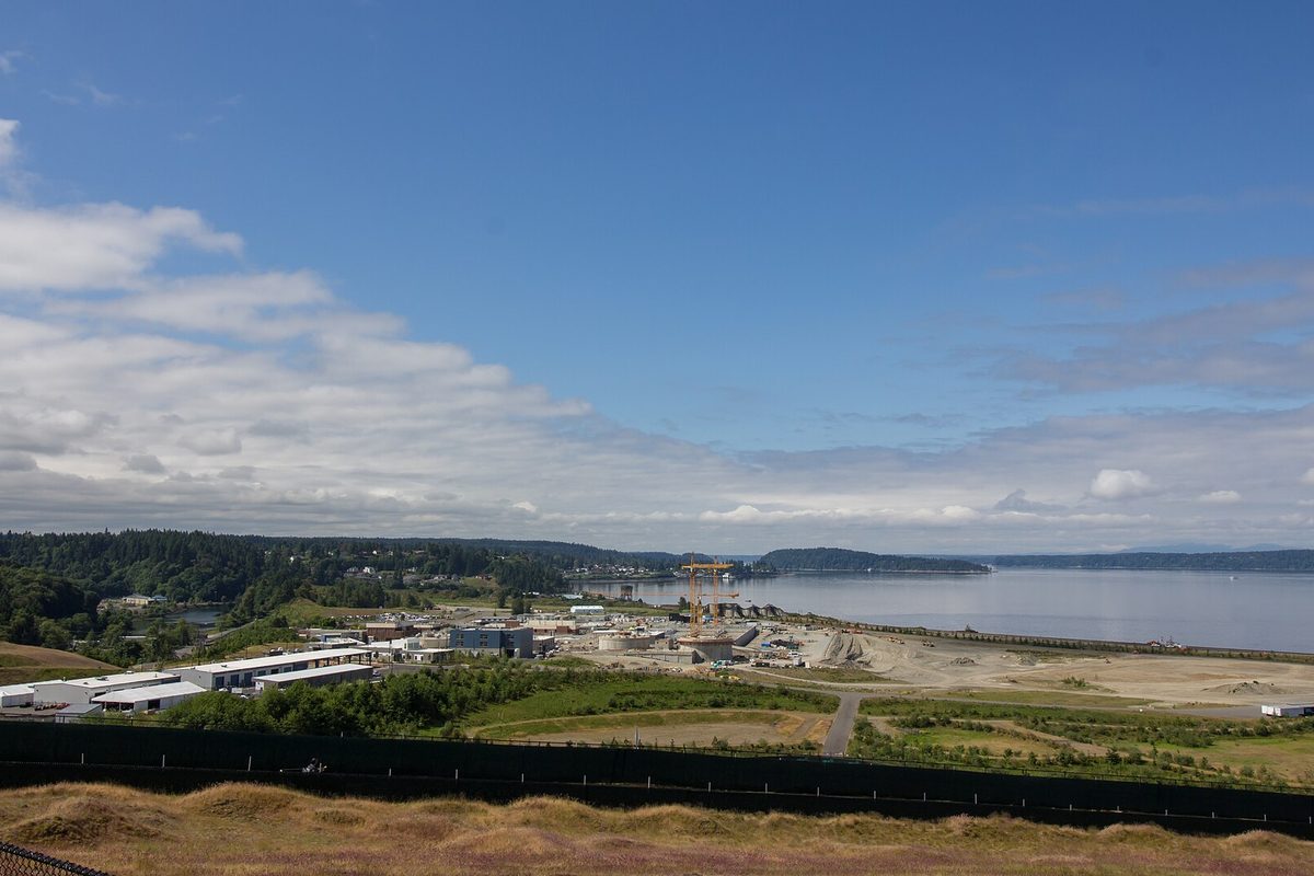 View over the Chambers Bay treatment plant and its sandy yards to Puget Sound and wooded islands under a blue sky with white clouds