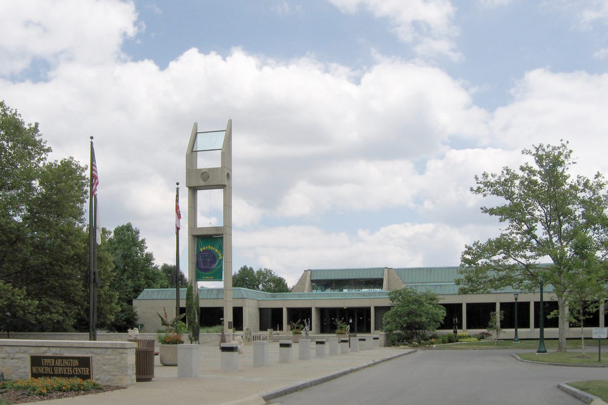 The Upper Arlington Municipal Services Center, a low building with a green roof, behind a tall open concrete tower, two flagpoles and a stone entrance sign