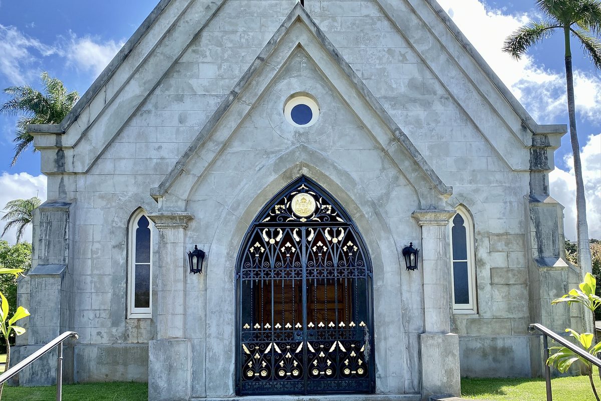 The gray stone Gothic chapel at Mauna Ala on Nuuanu Avenue, with an ornate black and gold iron gate in its pointed doorway, palm trees and blue sky
