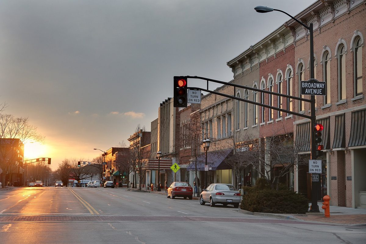 Sunset over East Main Street at Broadway Avenue in downtown Urbana, with brick storefronts, a red traffic light and parked cars