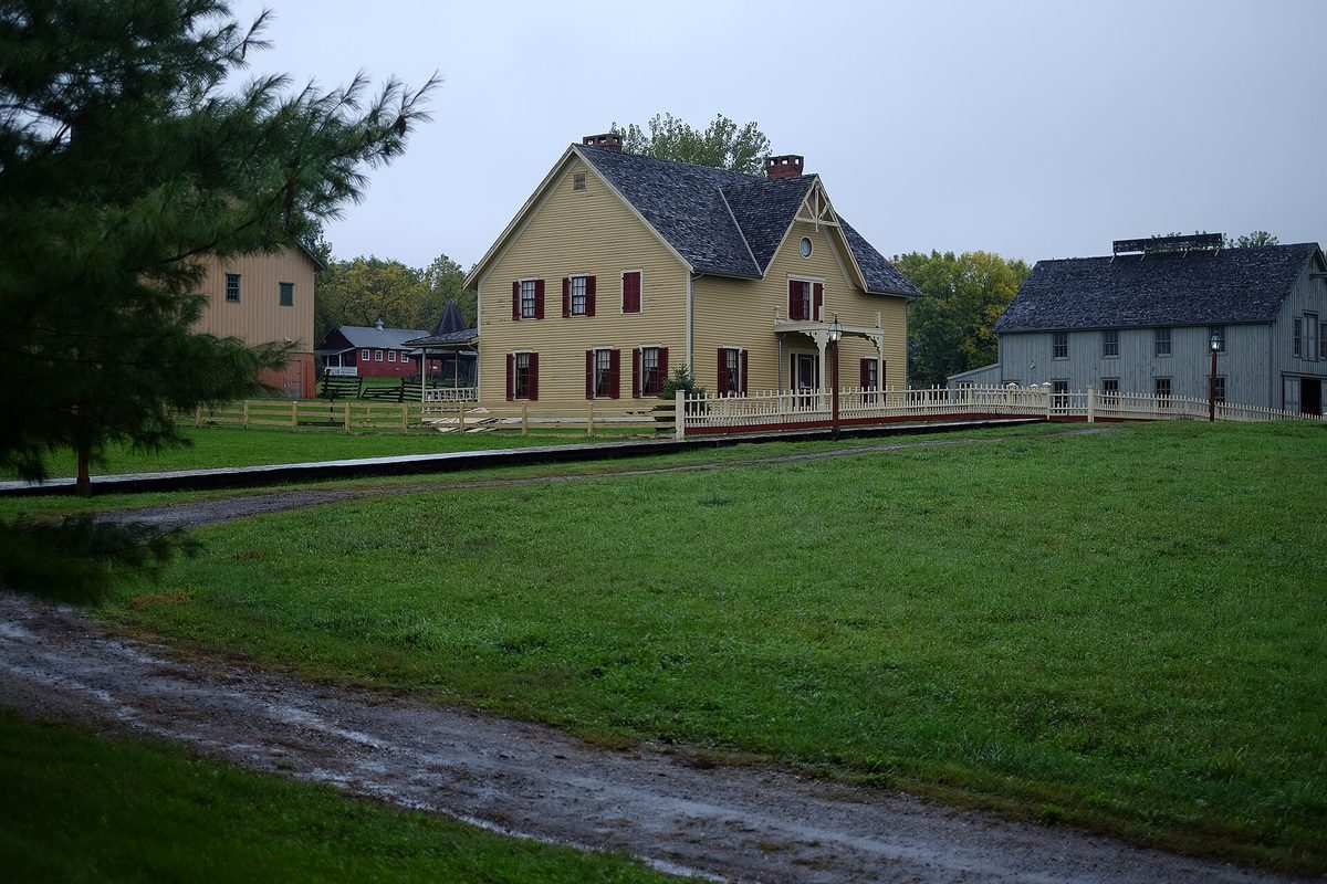 A yellow farmhouse with red shutters and a gray barn-like building beyond a green lawn and dirt path at Living History Farms in Urbandale, under an overcast sky