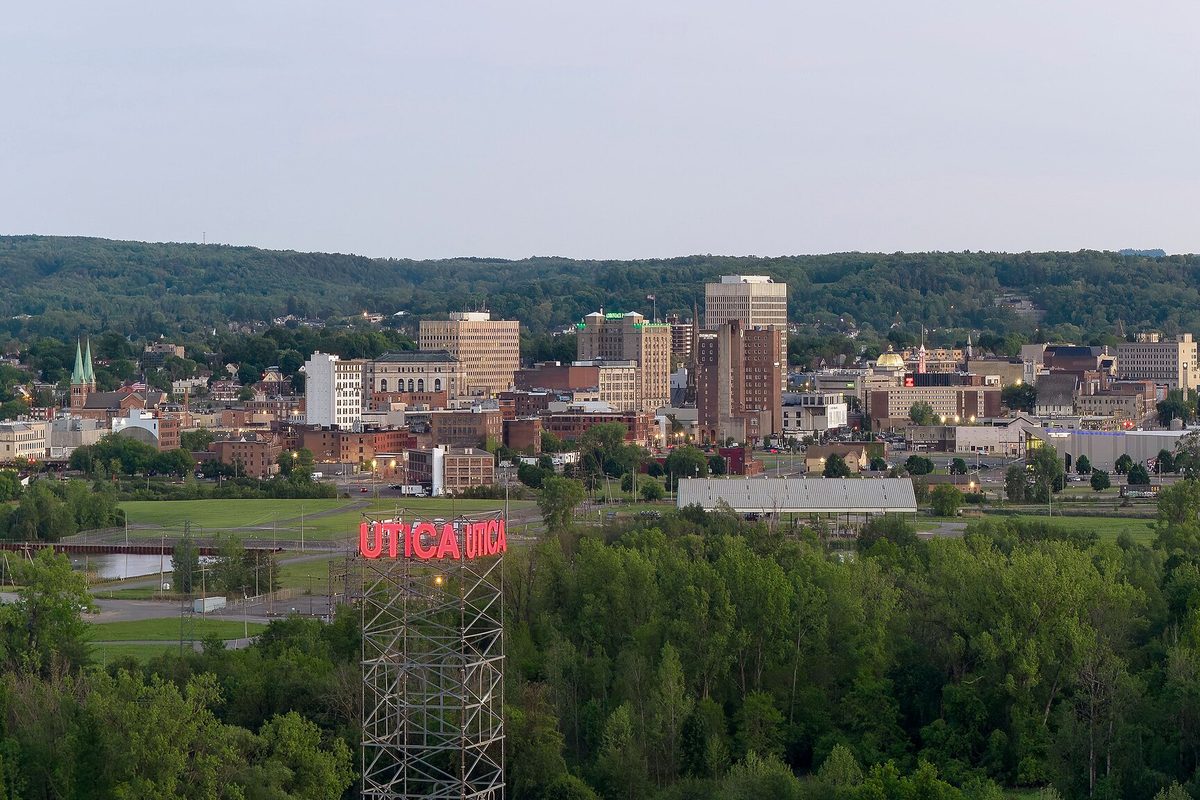 Downtown Utica at dusk seen across a band of trees, with a red UTICA sign lit on a steel tower in the foreground and wooded hills behind