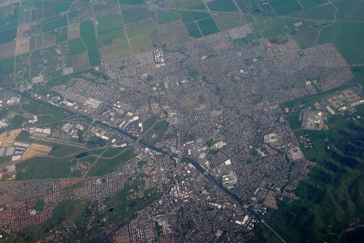 Aerial view of Vacaville: a grid of neighborhoods and a freeway cutting through, with green hills and farm fields at the edges