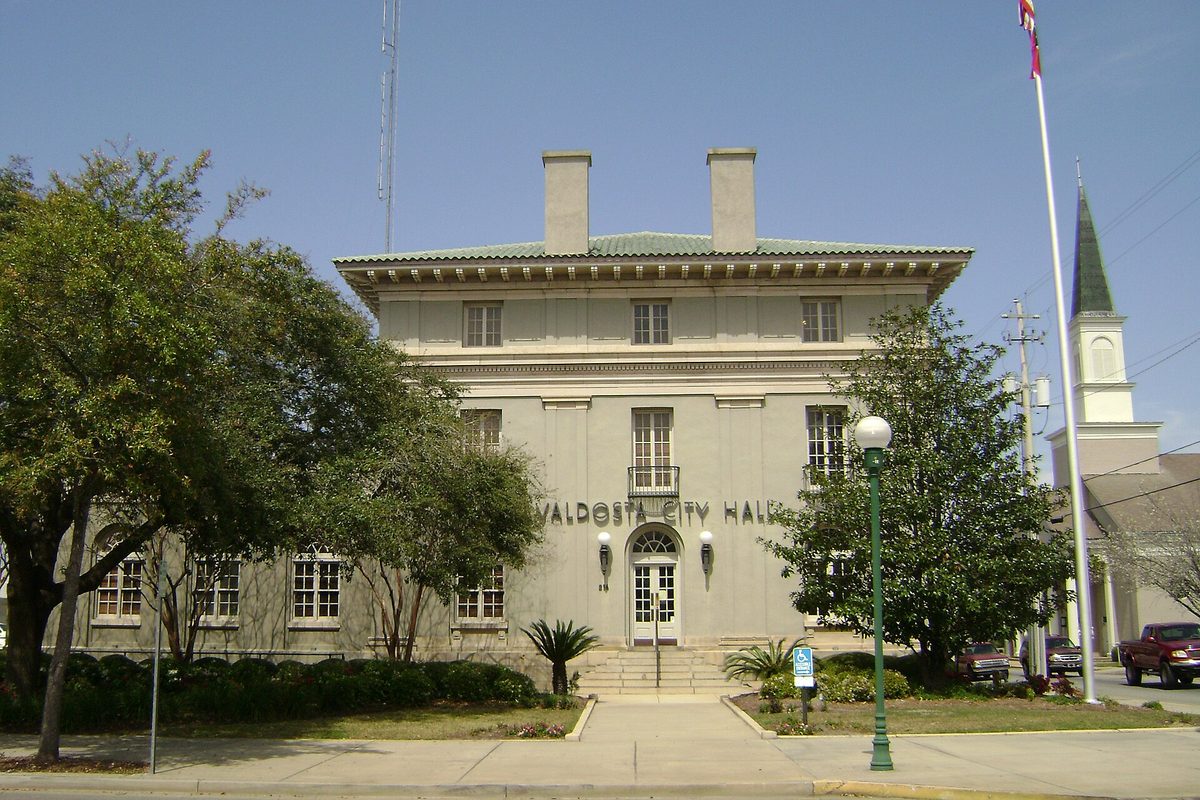 Valdosta City Hall, a pale two-story building with a green tile roof and two chimneys, framed by oak trees, small palms, a flagpole and a church steeple