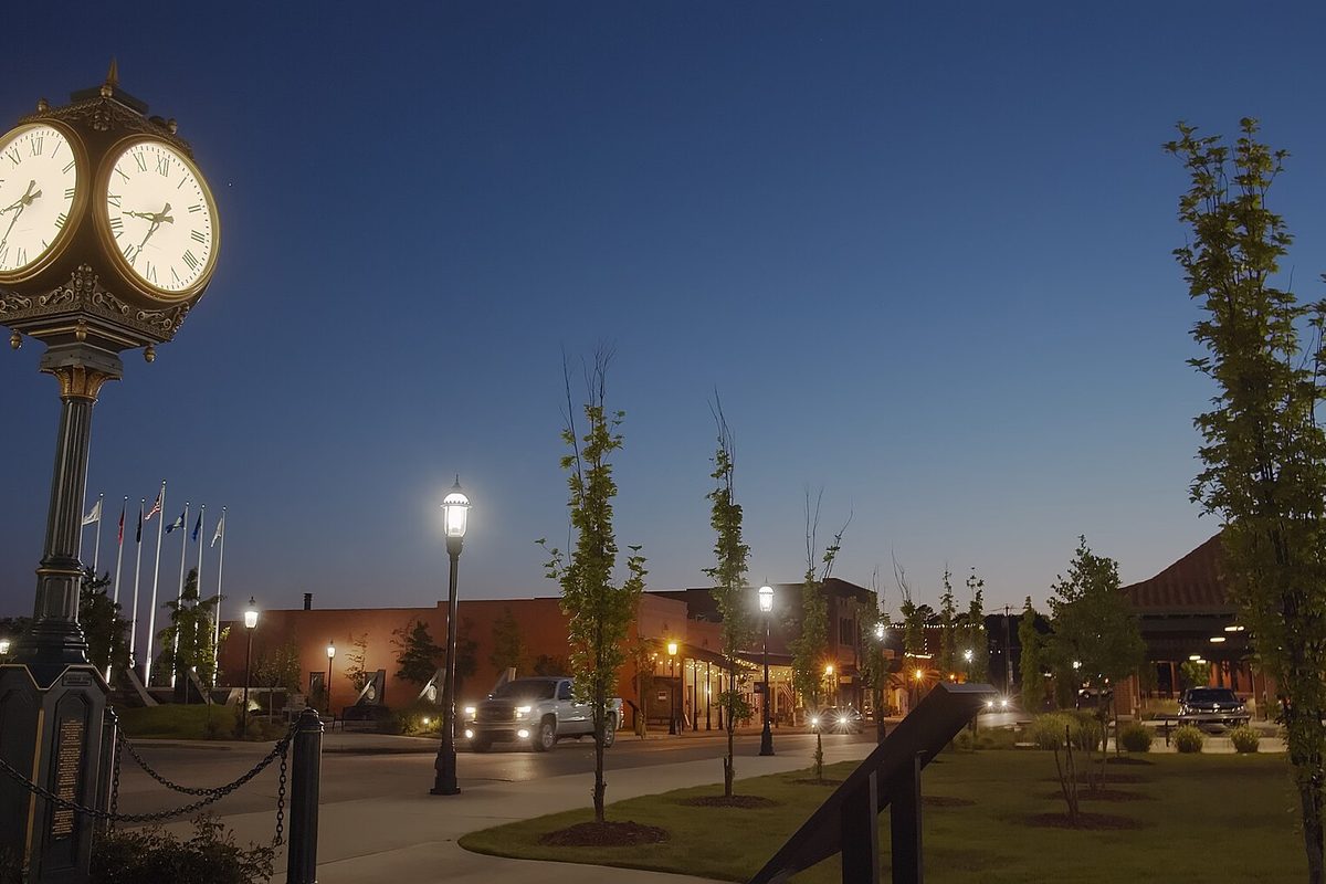 Freedom Park in downtown Van Buren at dusk, with a lit four-faced post clock, street lamps, flags, young trees and brick storefronts