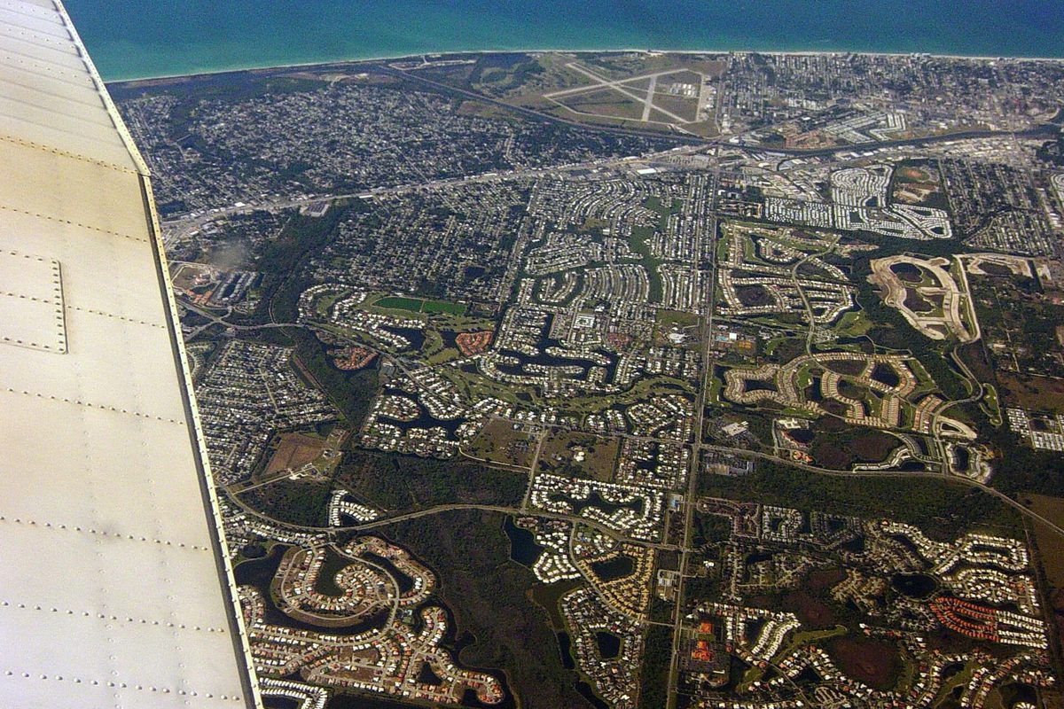 Aerial view from an airplane window over Venice, Florida, with the wing at left, curving neighborhoods and ponds below, an airport's runways and the Gulf beyond