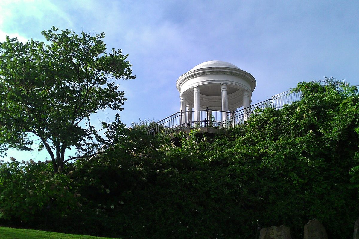 The white domed Sibyl Temple with its ring of columns on a hilltop above thick green trees and vines, under a blue sky