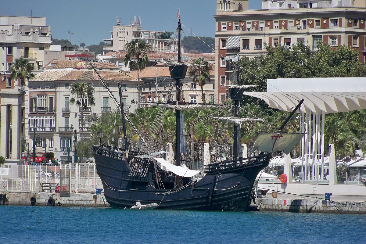 A replica of the sailing ship Nao Victoria, a dark wooden galleon with three masts, moored at a harbor quay with palm trees and apartment buildings behind it