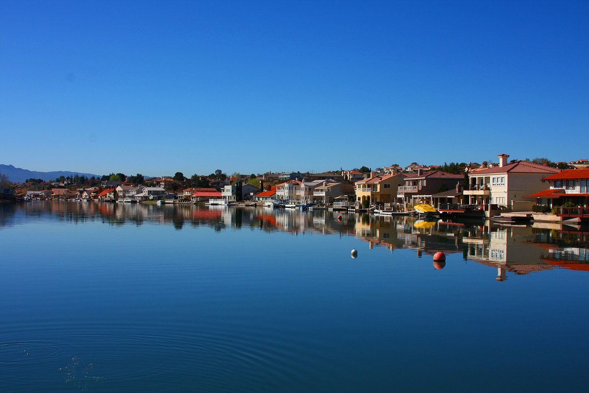 Lakefront homes with red tile roofs reflected in the still water of Spring Valley Lake in the Victor Valley, under a clear blue sky