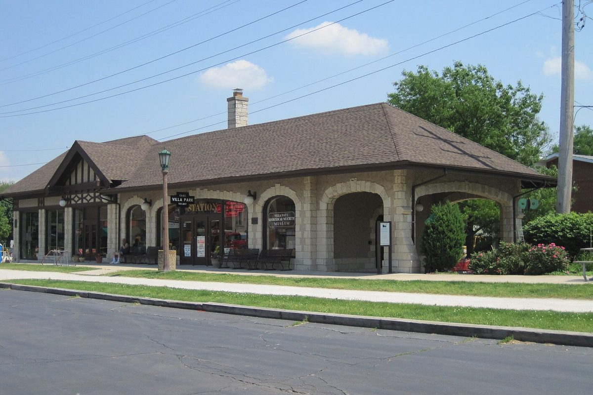The 1929 Villa Avenue train station in Villa Park, a stone building with arched openings, a brown hip roof and a chimney, beside a quiet street on a sunny day