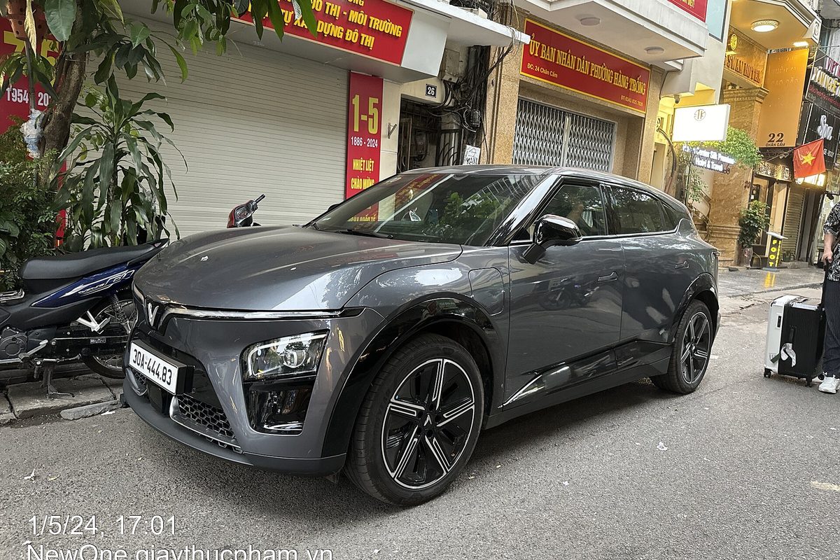 A gray VinFast VF 7 crossover parked on a city street in front of shops, with a motorbike beside it