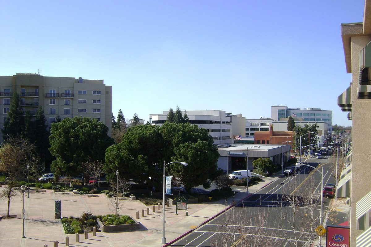 Acequia Avenue in downtown Visalia seen from above, with office buildings, a tree-lined plaza and a wide street under a clear sky