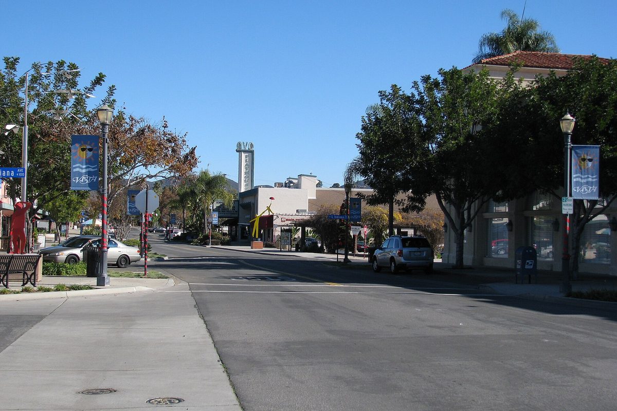 Looking east down Main Street in downtown Vista on a clear day, with blue street banners on lamp posts, shade trees, a palm and parked cars