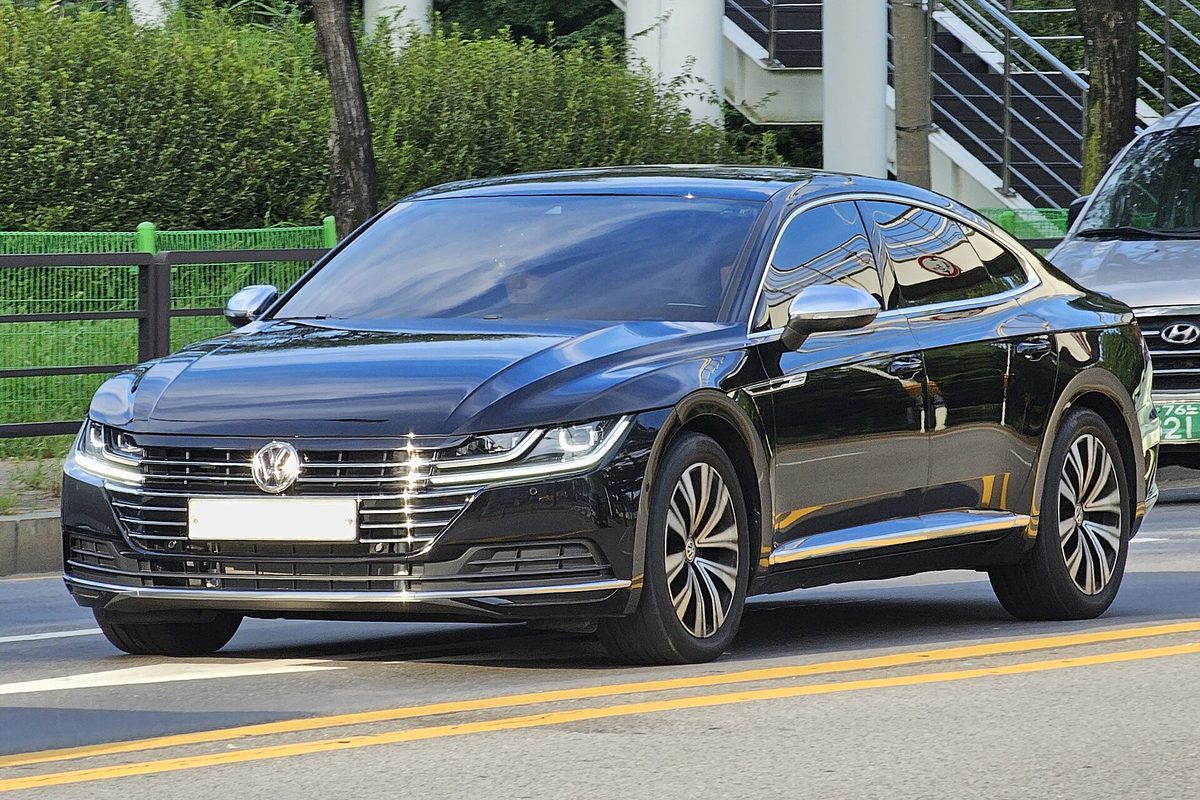 A black Volkswagen Arteon liftback driving on a city road past a green fence and trees