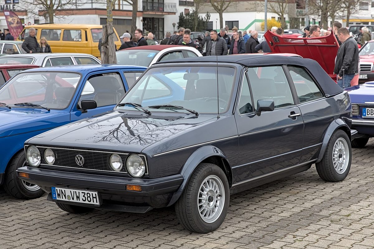 A dark gray first-generation Volkswagen Golf Cabriolet with its black soft top up, parked on cobblestones at an outdoor car meet