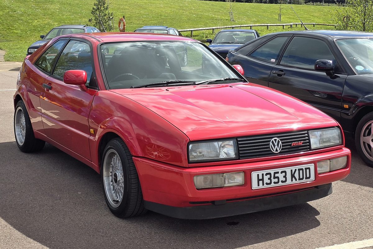 A red Volkswagen Corrado coupe parked in a lot next to a black Volkswagen, with a grassy hill behind