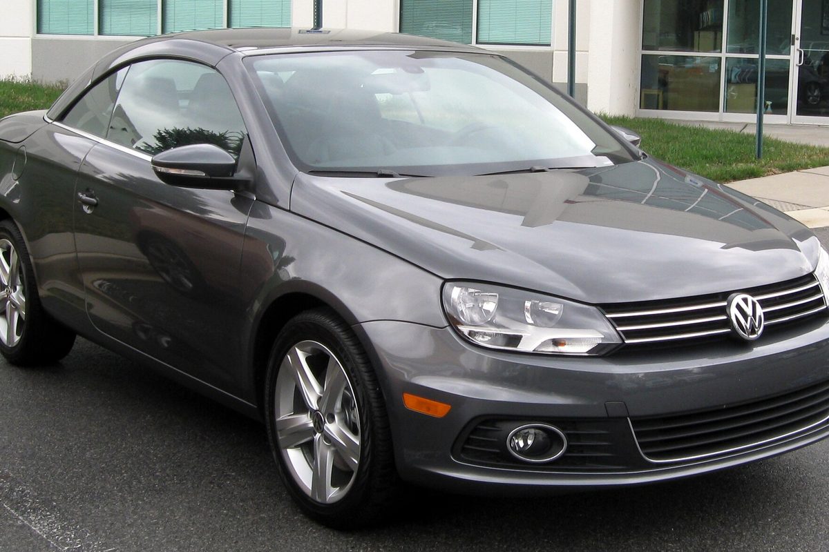 A dark gray Volkswagen Eos two-door with its roof closed, parked on dark pavement beside a lawn and a glass-front building