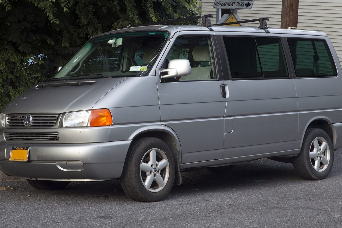 A silver Volkswagen Eurovan passenger van with a roof rack, parked on a street in front of trees and a white building