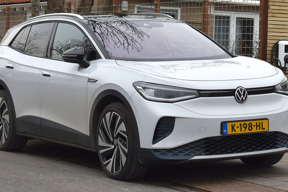 A white Volkswagen ID.4 electric crossover with a Dutch license plate, parked on a brick street beside a fence and a wall charger