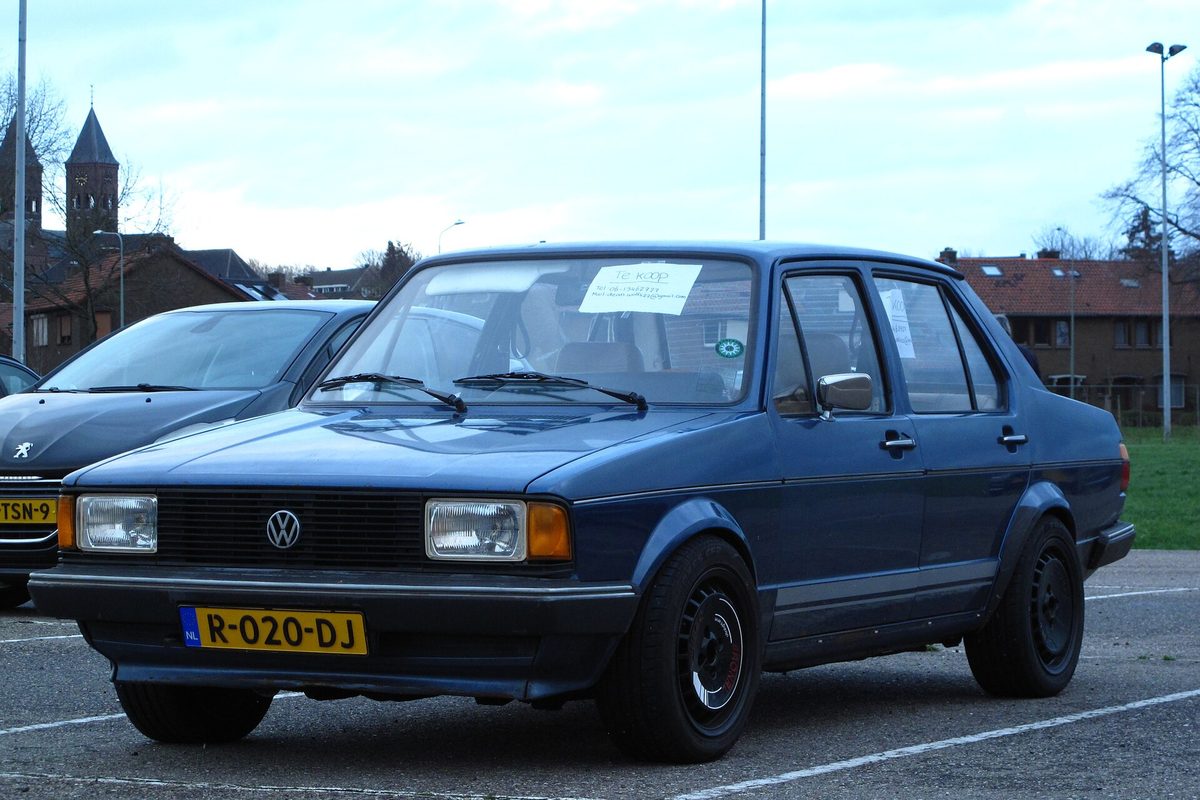 A boxy dark blue early Volkswagen Jetta sedan with black steel wheels and a paper sign behind the windshield, parked in a lot