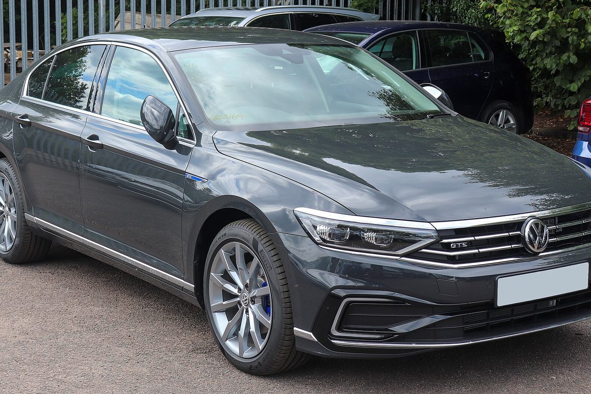 A dark gray Volkswagen Passat GTE sedan with silver alloy wheels, parked in front of a metal fence and hedges