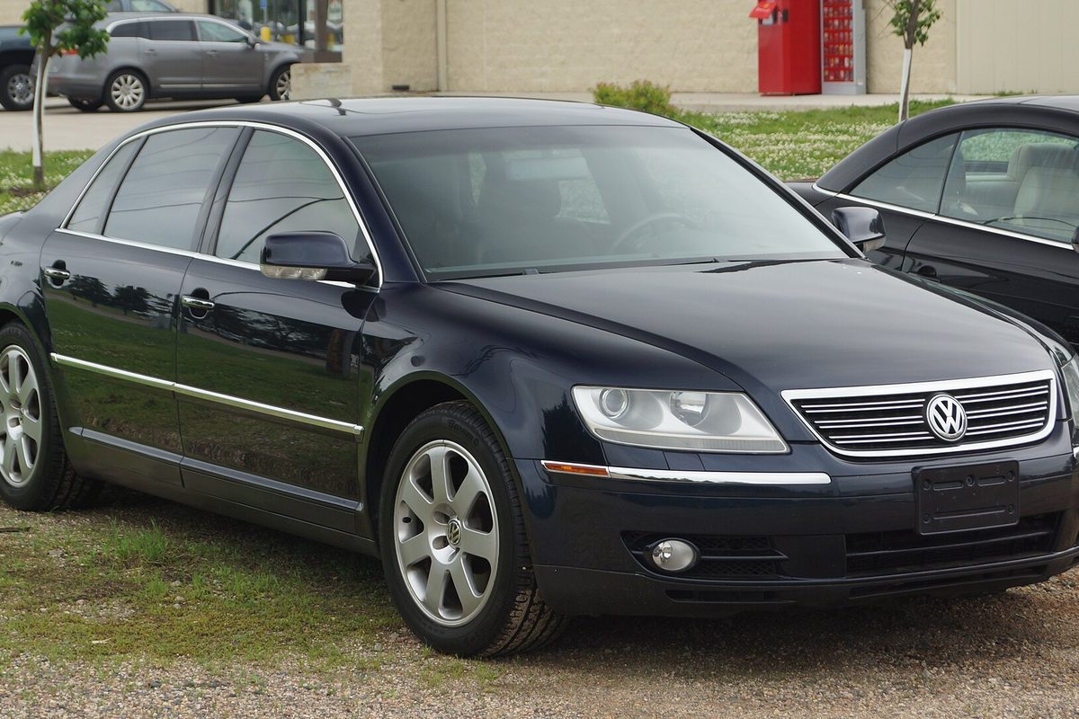 A dark blue 2004 Volkswagen Phaeton sedan with chrome trim, parked on gravel at the edge of a grassy lot