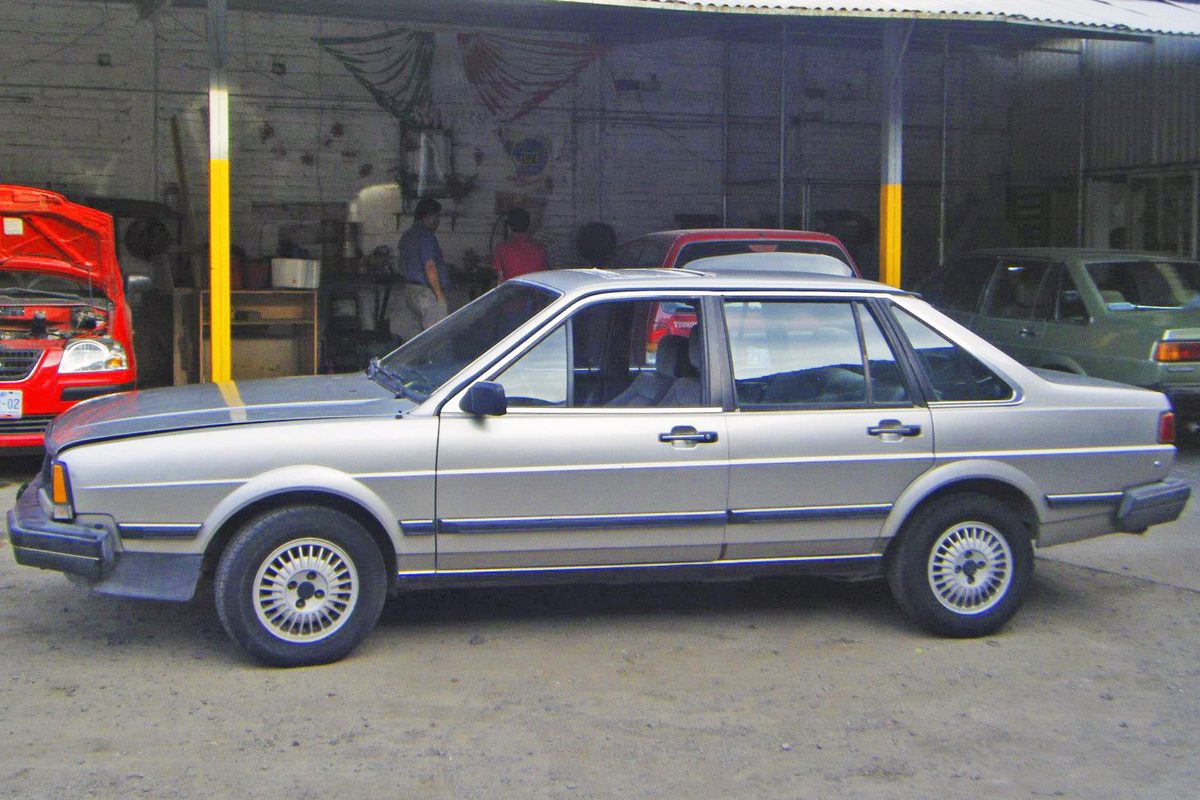 A silver-beige 1985 Volkswagen Quantum GL5 four-door sedan parked in a workshop yard beside other cars