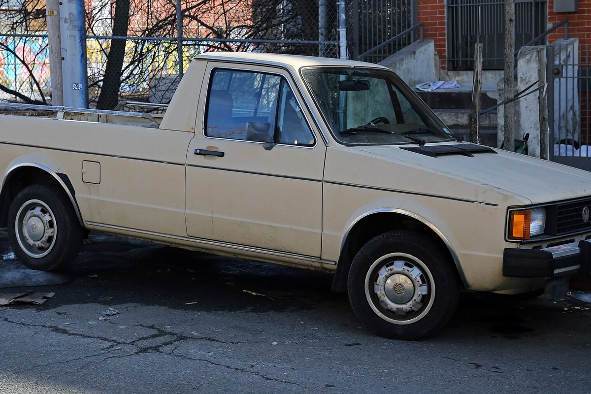 A faded tan Volkswagen Rabbit pickup parked on a city street beside a chain-link fence and a brick building