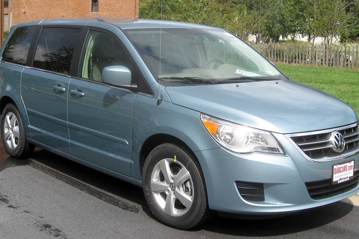 A light blue Volkswagen Routan minivan with a dealer sticker on the windshield, parked on asphalt beside a lawn and a wooden fence