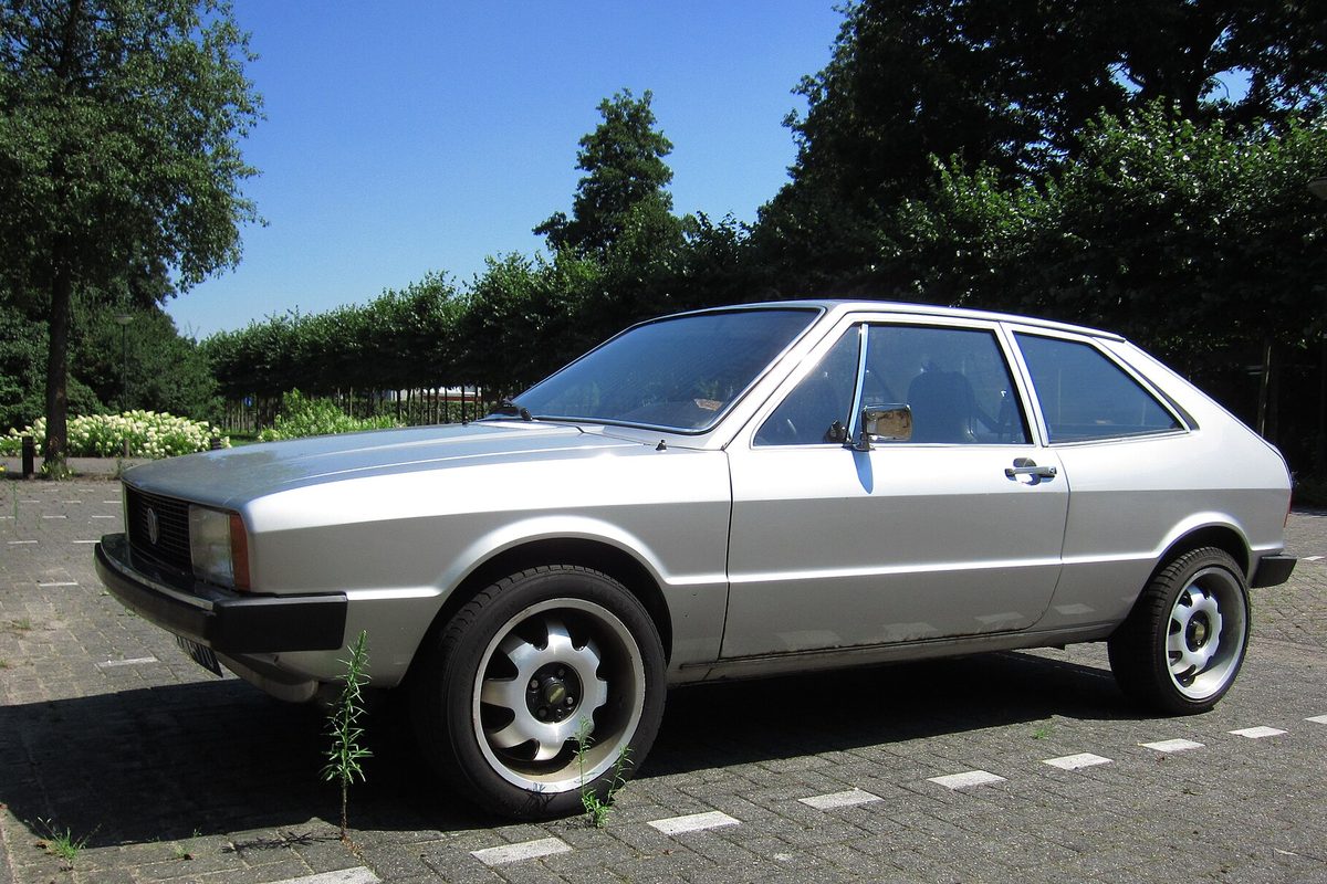 A silver first-generation Volkswagen Scirocco hatchback coupe with aftermarket alloy wheels, parked on brick pavers by a hedge