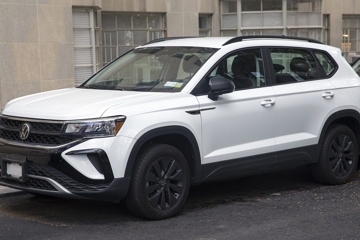 A white Volkswagen Taos small SUV with black wheels and a black roof rail, parked at a curb in front of a stone office building