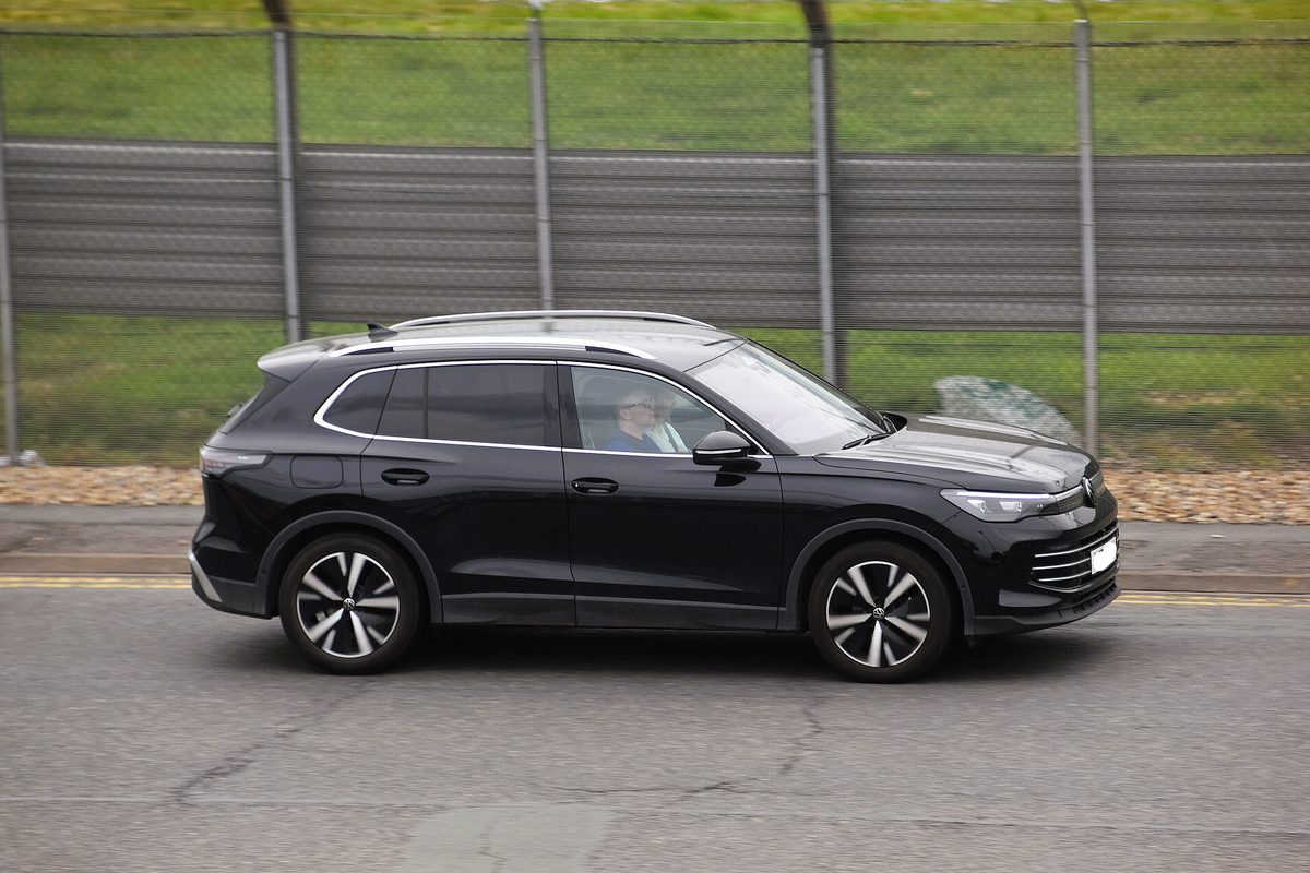 A black 2024 Volkswagen Tiguan SUV seen from the side, driving past a tall metal fence