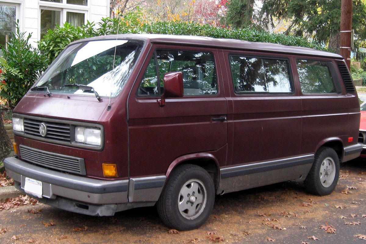 A maroon Volkswagen Vanagon with gray bumpers parked on a leafy residential street in autumn