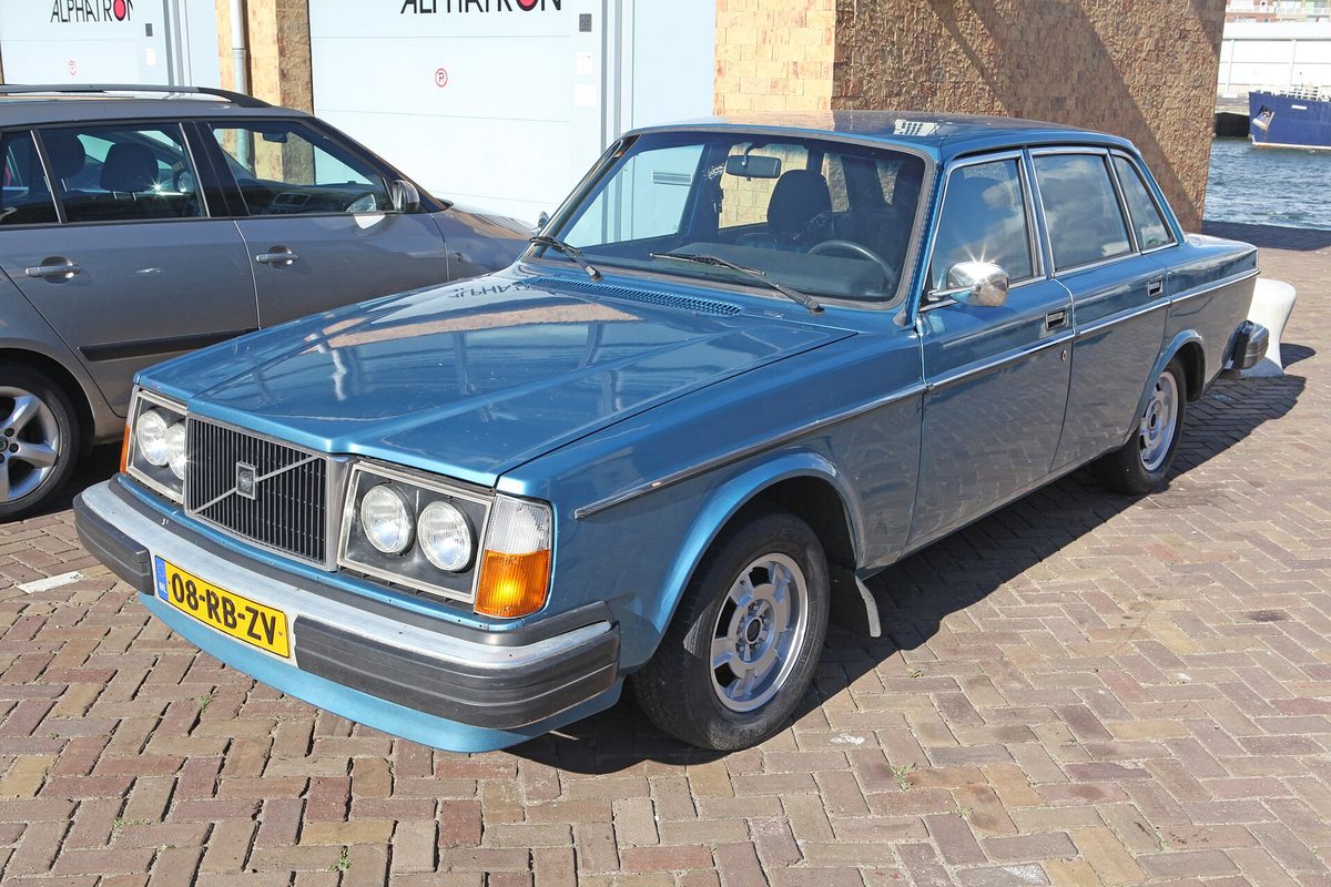 A light blue 1978 Volvo 240 sedan with round headlights, parked on brick pavement by a harbor