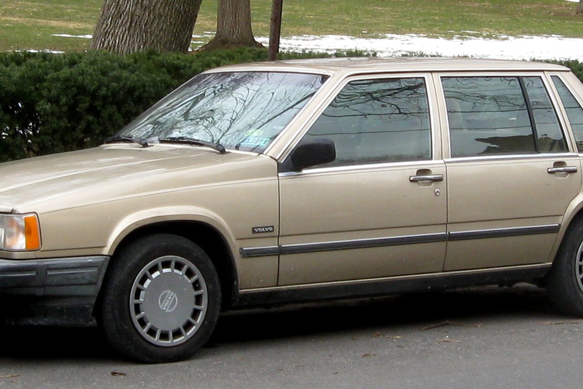 A tan Volvo 700 Series sedan with gray bumpers and flat gray hubcaps, parked on a street beside a hedge