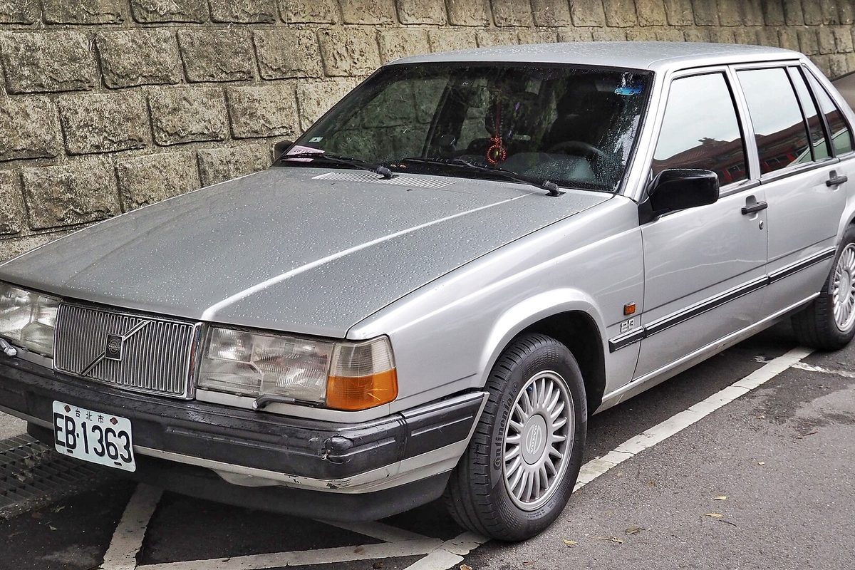 A silver 1990 Volvo 940 sedan parked in front of a gray stone wall