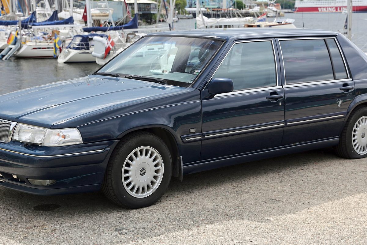A dark blue 1995 Volvo 960 Executive sedan parked by a marina, with sailboats and a ferry behind it