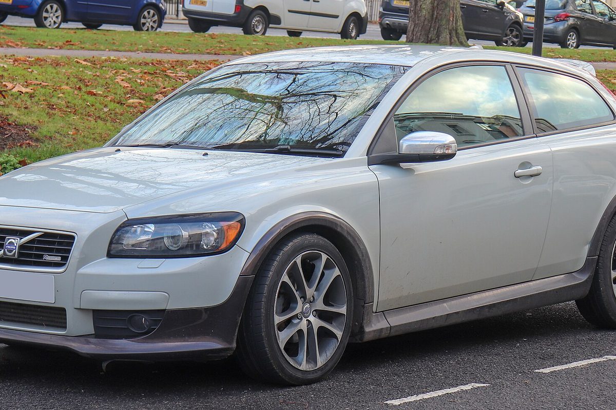 A pale silver-green Volvo C30 hatchback parked at a curb beside a lawn covered in fallen leaves
