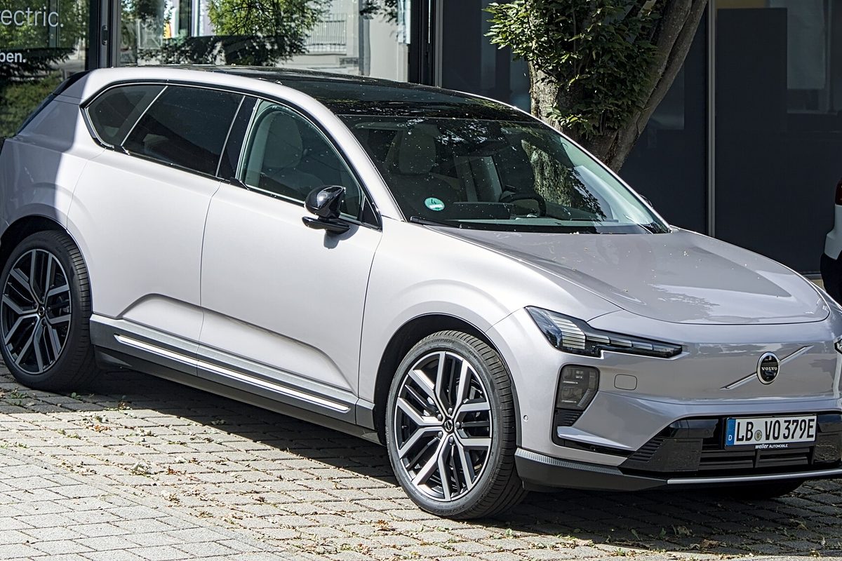 A light silver Volvo EX60 electric SUV with large two-tone wheels, parked on paving stones in front of a glass showroom