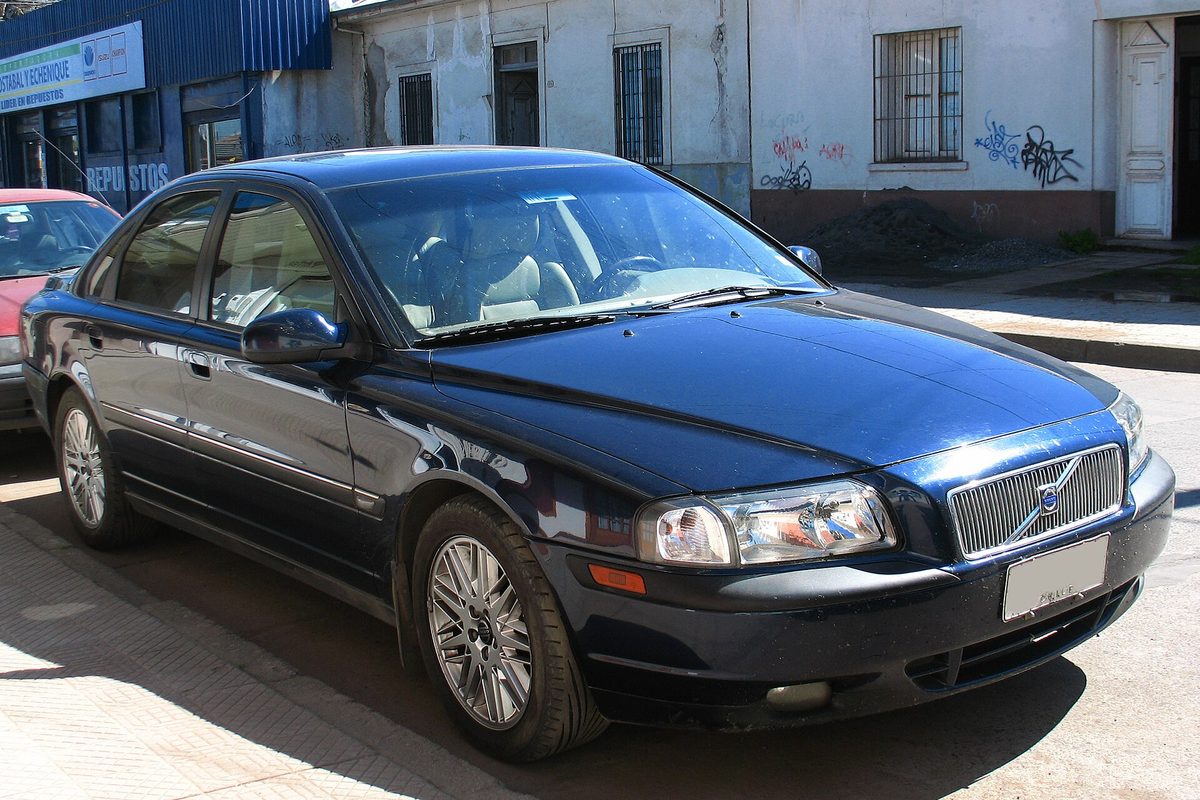 A dark blue first-generation Volvo S80 sedan parked on a street in front of a white building