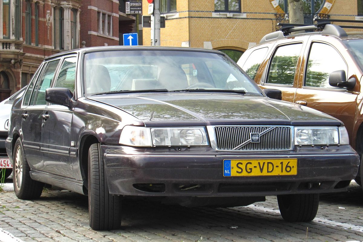 A dark plum late-1990s Volvo S90 sedan parked on a cobblestone street in front of brick buildings