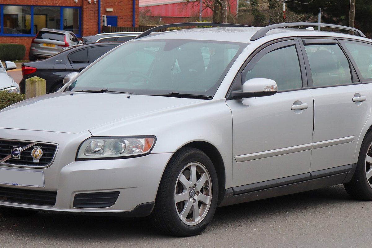 A silver Volvo V50 station wagon with roof rails parked in a lot in front of a red brick building