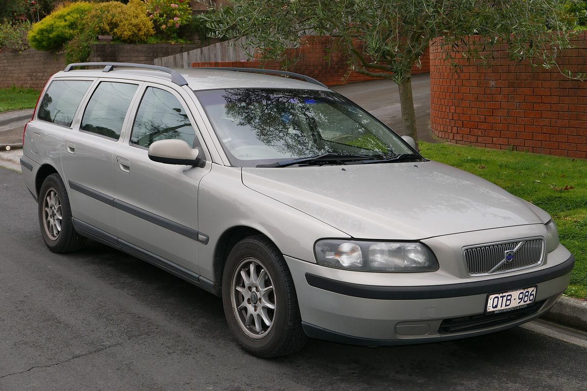 A silver 2001 Volvo V70 station wagon parked at a curb beside a brick wall and a garden