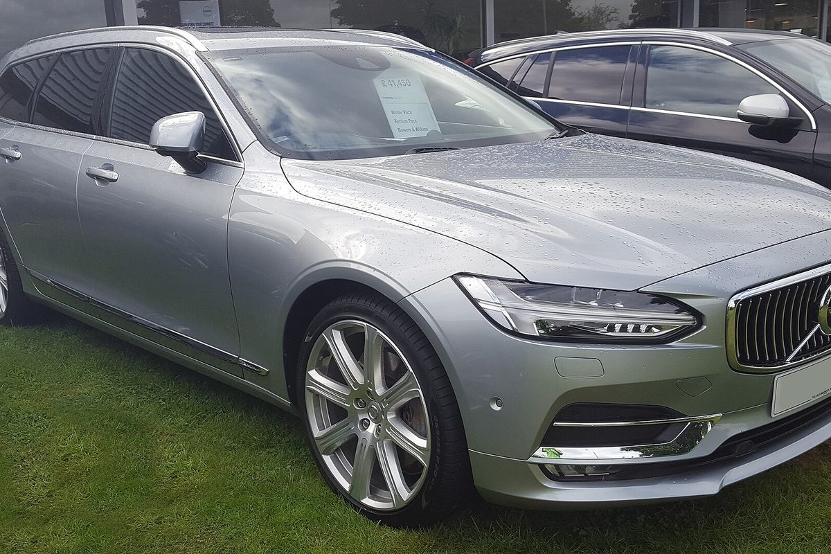 A silver Volvo V90 wagon parked on wet grass outside a showroom, a printed card behind the windshield