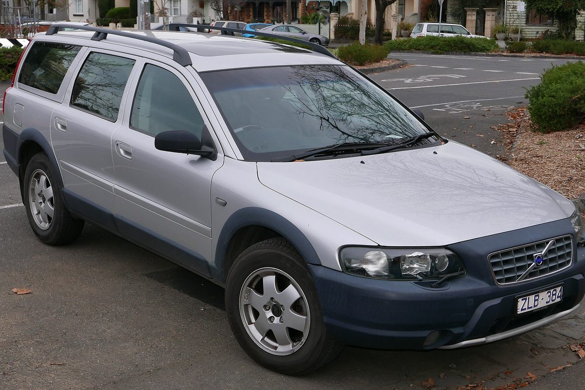A silver Volvo XC70 wagon with dark gray lower body cladding, roof rails and silver alloy wheels, parked at a curb