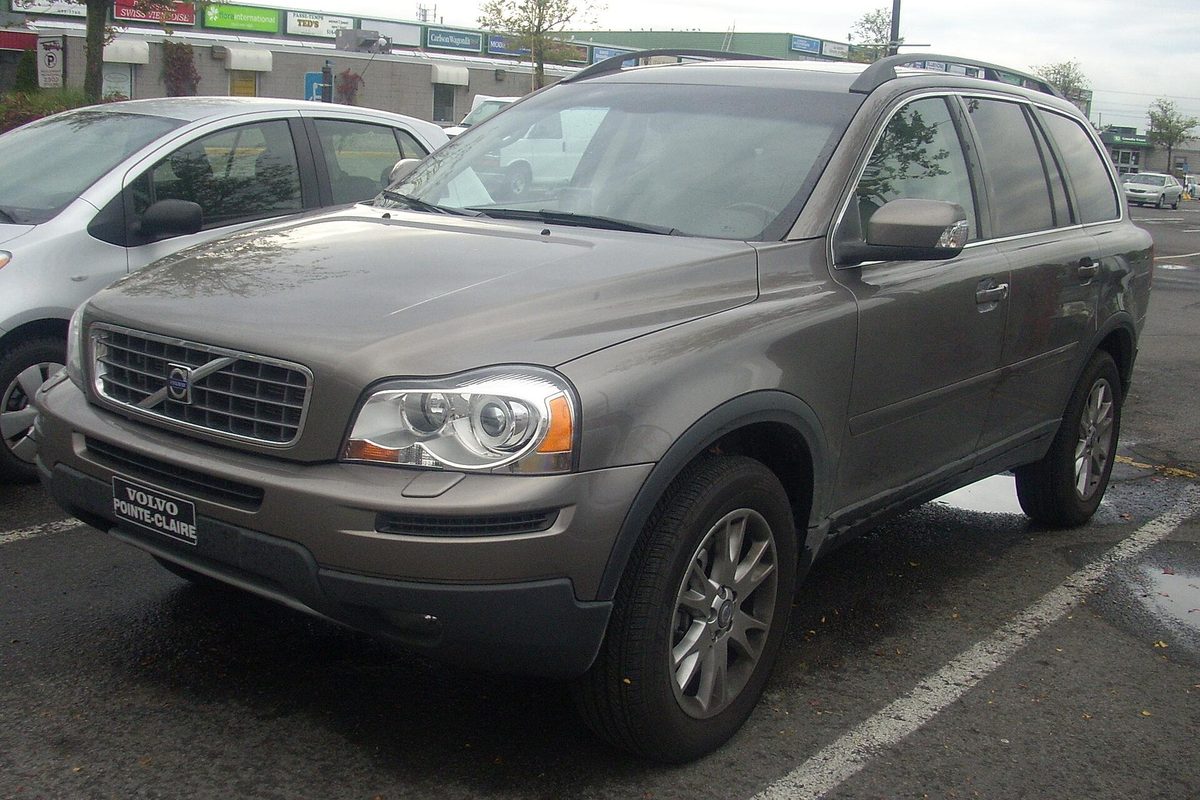 A bronze first-generation Volvo XC90 SUV parked in a wet shopping-center lot, seen from the front