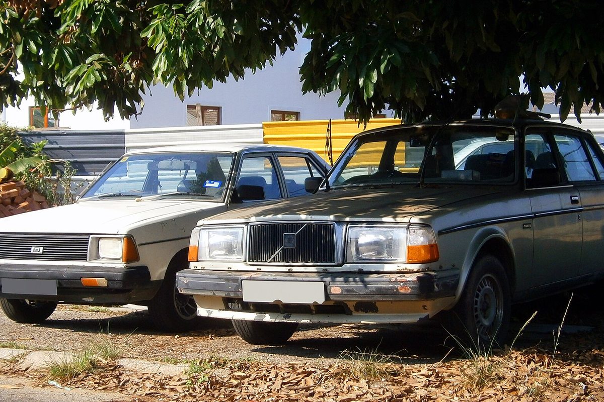 A weathered tan 1986 Volvo 240 sedan parked on dirt and dry leaves under a tree, beside a white 1981 Datsun 120Y, in Ipoh, Malaysia