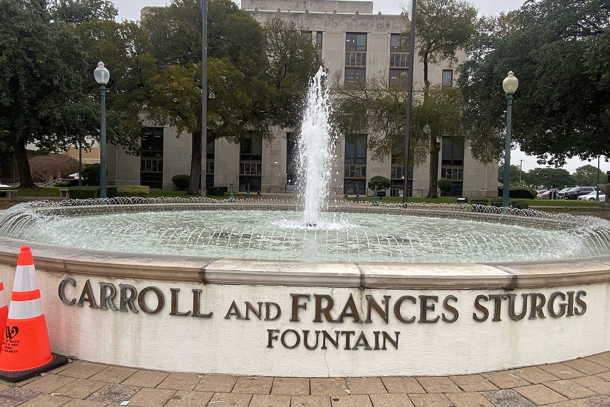 The round Carroll and Frances Sturgis Fountain spraying water in front of Waco City Hall, with lamp posts, trees and an orange traffic cone at the edge