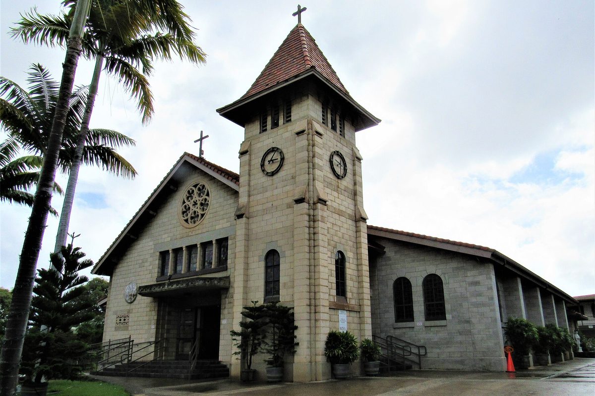 Saint Joseph Catholic Church in Waipahu, a stone church with a square clock tower and red tile roof, framed by palm trees under a cloudy sky
